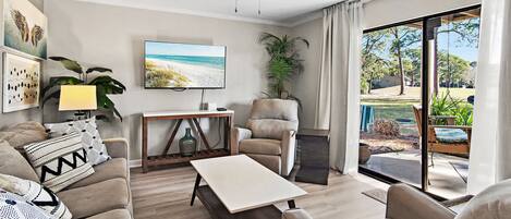 A living room with a sofa, armchair, coffee table, TV, ceiling fan, and a view of the outdoors.