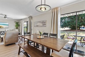 A dining area with a wooden table, benches, and chairs. There is a ceiling fan, a painting on the wall, and a window with curtains. - 🏖️Ground Floor 2BR | Pet Friendly | Pools + Golf Course | Seascape 91B🌞 (Miramar Beach)