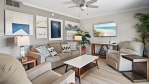 A modern living room with a sofa, armchairs, a coffee table, and a TV. There are wall art pieces and a ceiling fan. - 🏖️Ground Floor 2BR | Pet Friendly | Pools + Golf Course | Seascape 91B🌞 (Miramar Beach)