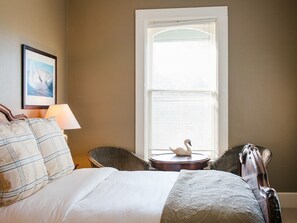 A bedroom with a bed, two wicker chairs, a round table with a swan decoration, a lamp, and a framed picture on the wall.