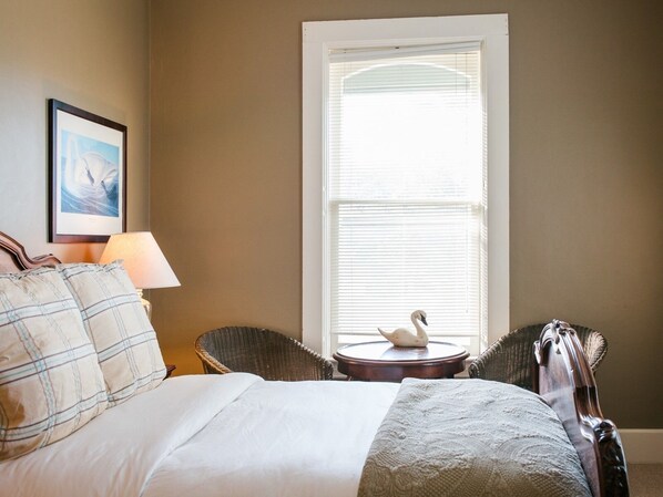 A bedroom with a bed, two wicker chairs, a round table with a swan decoration, a lamp, and a framed picture on the wall.