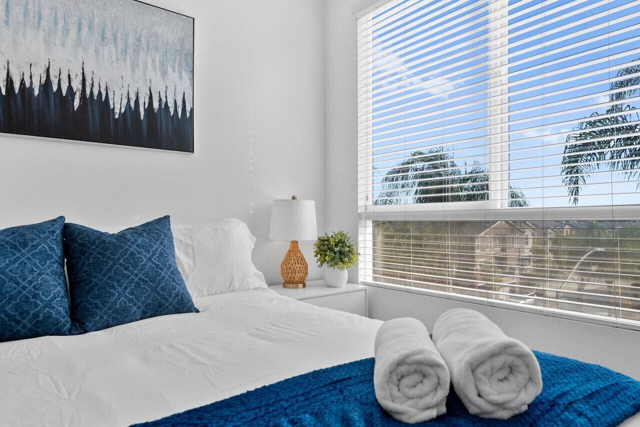 A hotel room with a large bed, white bedding, blue pillows, a blue throw blanket, a white lamp, a small plant, and a window with blinds.