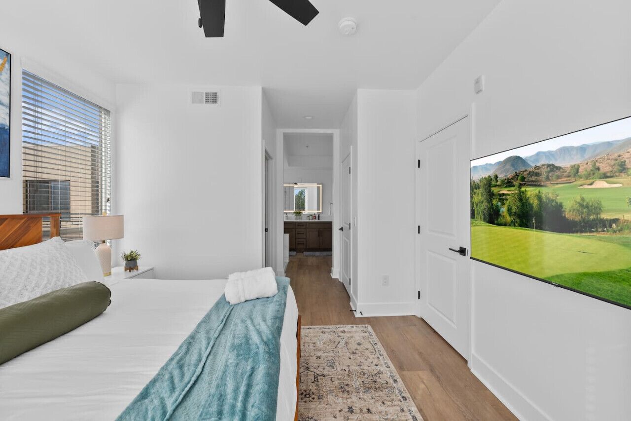 A bedroom with a bed, a nightstand, a lamp, a window with blinds, a view of a golf course, and a door leading to a bathroom.