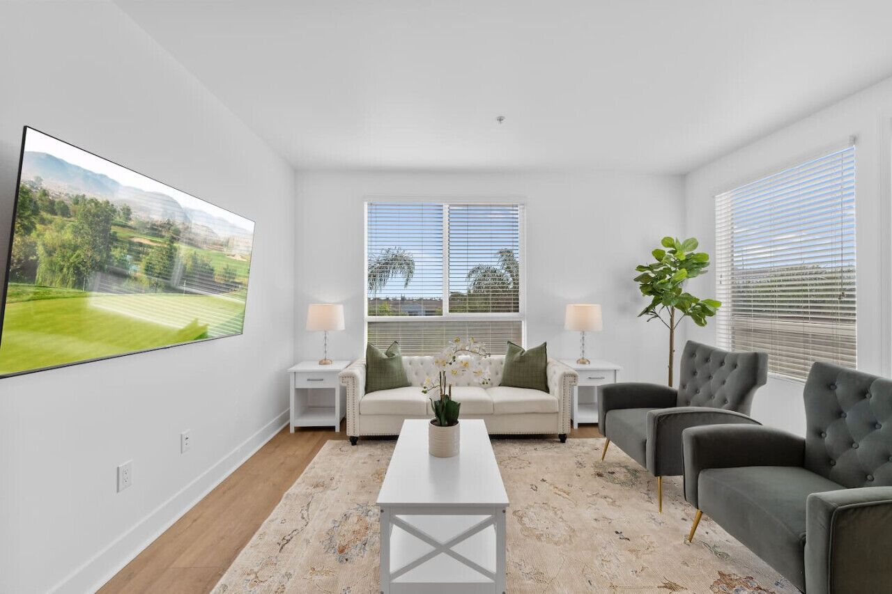 Un salon moderne avec un grand téléviseur à écran plat, un canapé blanc, un fauteuil gris et une table basse. On y trouve deux tables de chevet avec des lampes et une plante en pot.