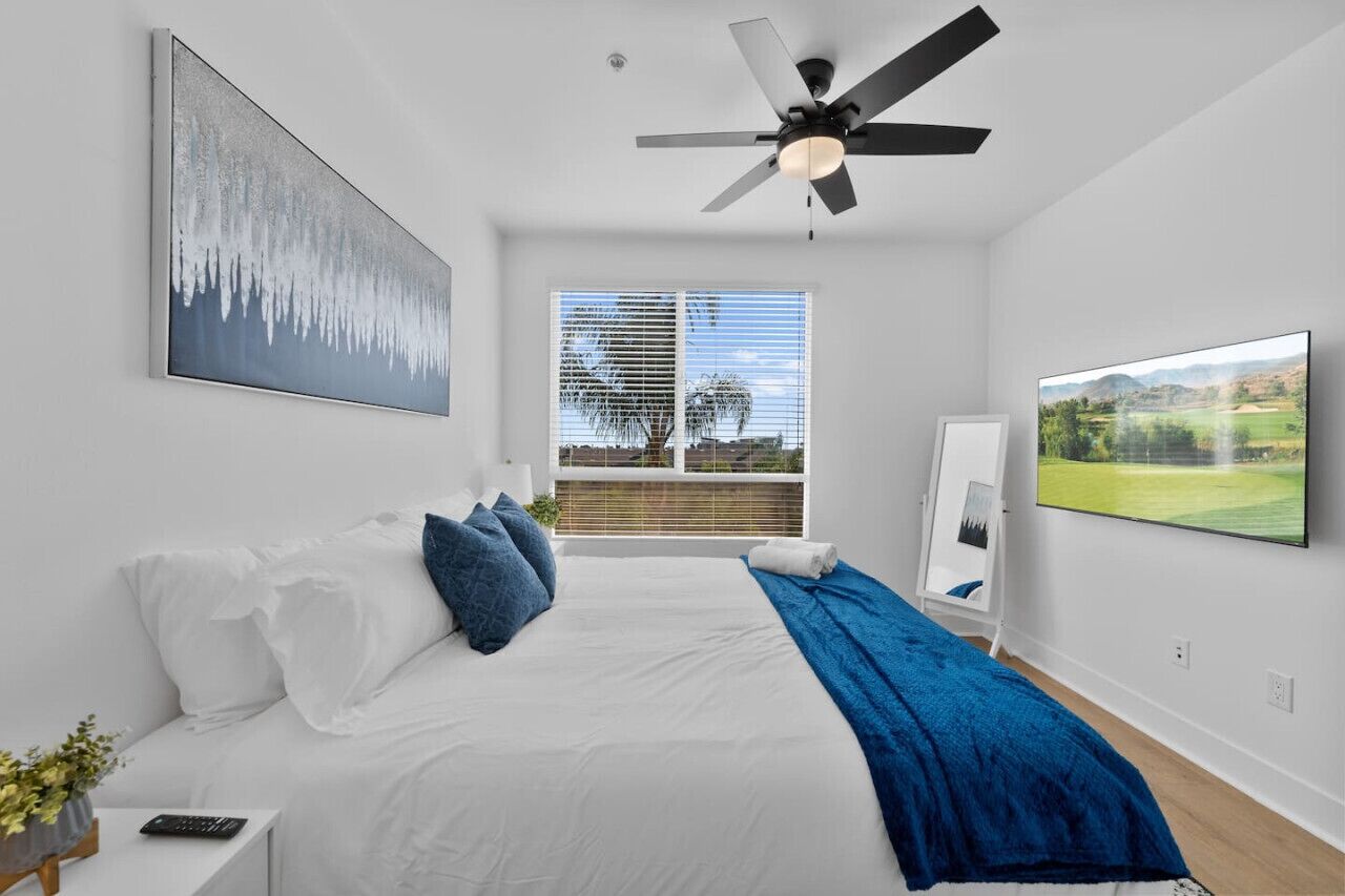 Ein Schlafzimmer mit einem Bett, einem Deckenventilator, einem großen Fenster mit Jalousien und einem Fernseher an der Wand.