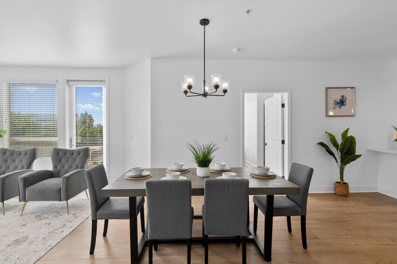 Un comedor con una mesa rectangular, sillas tapizadas en gris, una araña y una planta en maceta.