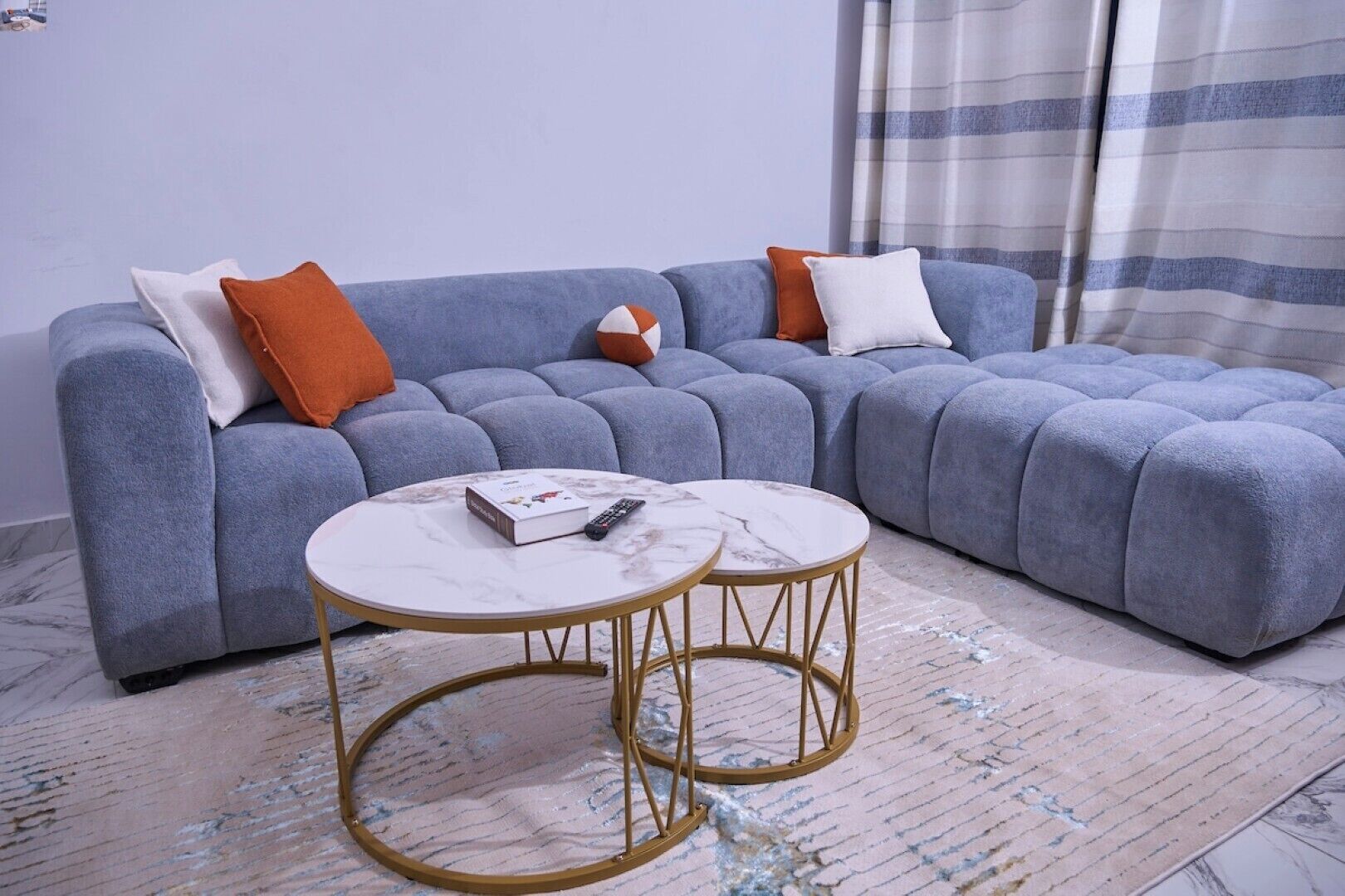 Ein modernes Wohnzimmer mit einem grauen Ecksofa, zwei runden Goldtischen und einem gestreiften Vorhang im Hintergrund.