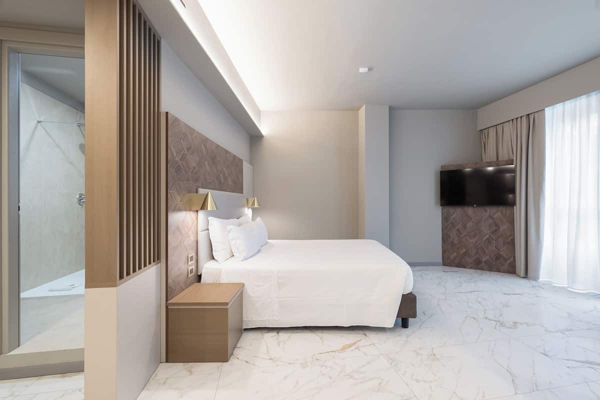 A modern hotel room with a large bed, a bedside table, a TV, and a glass shower area.