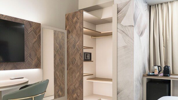 Ein modernes Hotelzimmer mit Flachbildfernseher, Essbereich mit grünem Sessel, integriertem Regal, Kaffeemaschine und kleinem Kühlschrank.