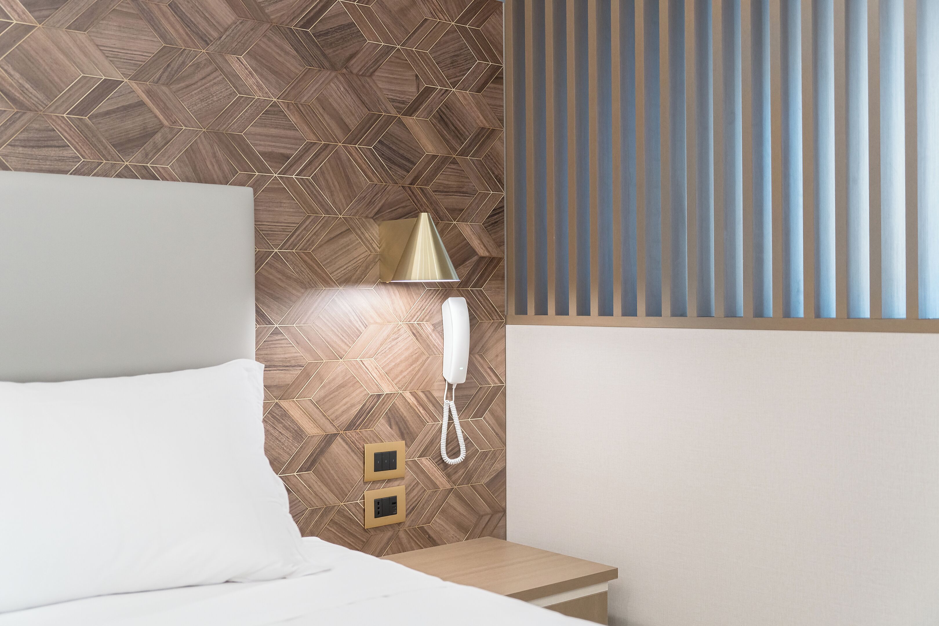 Ett hotellrum med en vägg som har ett träpanelmönster i fiskbensstil, en sänglampa, en telefon och ett fönster med vertikala persienner.