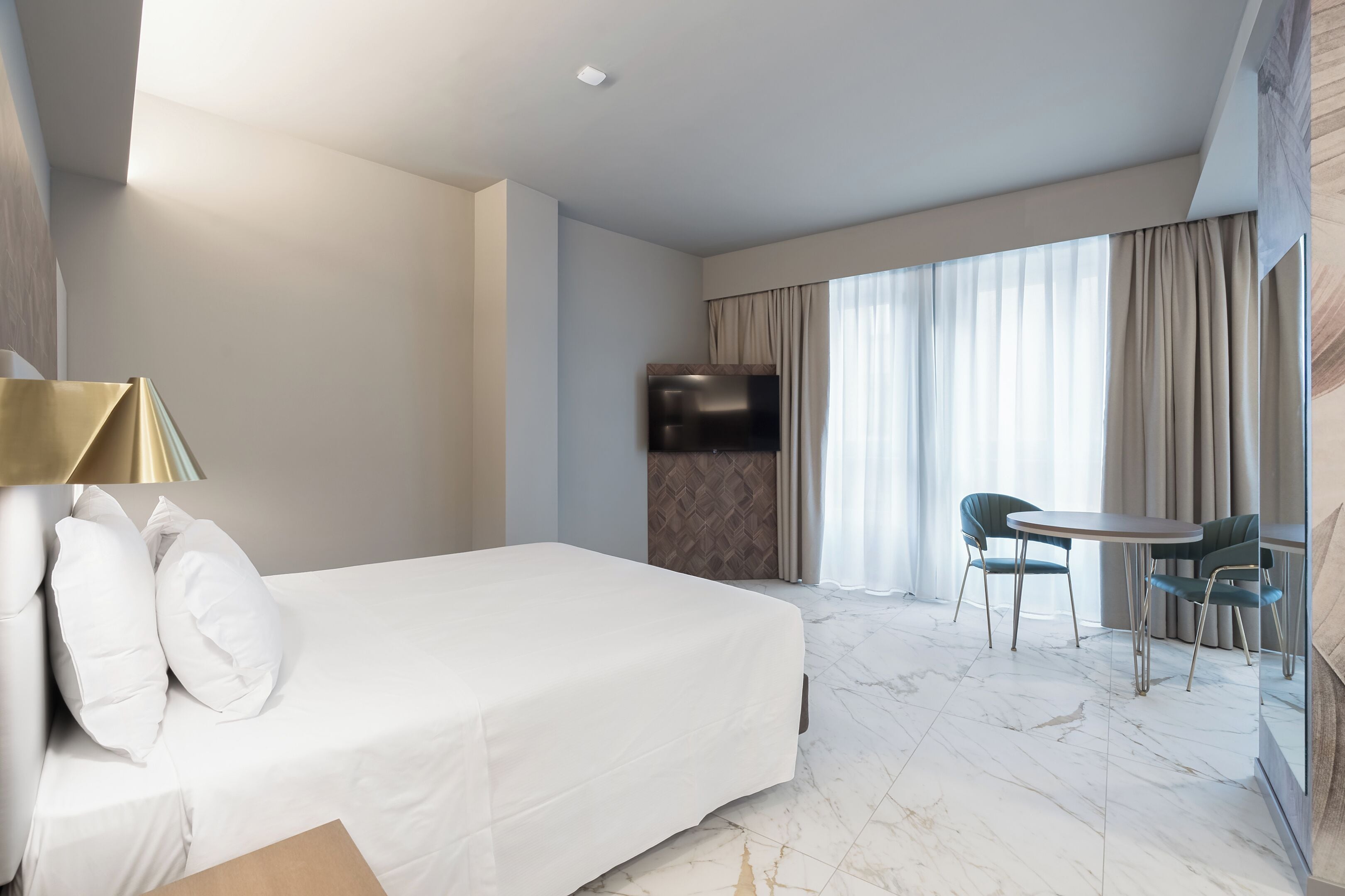A hotel room with a bed, a TV, a dining table with chairs, and a large window with curtains.