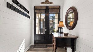 A hallway with a black door, a coat rack with hooks, a round mirror, and a table with a lamp. - Steps from the sand! 4BR/3BA private home w/ heated pool, 6 seater gas golf cart (Panama City Beach, FL)