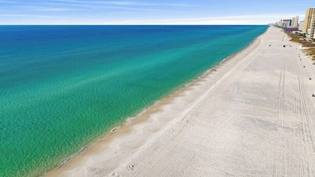 A beach with clear turquoise water, a sandy shore, and buildings along the coastline. - Steps from the sand! 4BR/3BA private home w/ heated pool, 6 seater gas golf cart (Panama City Beach, FL)