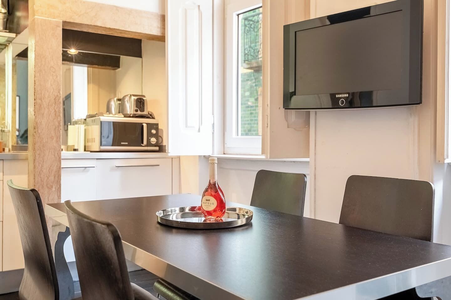 Et moderne køkken med et spisebord, stole, en flaske Mateus vin, en mikroovn og et fladskærms-TV.