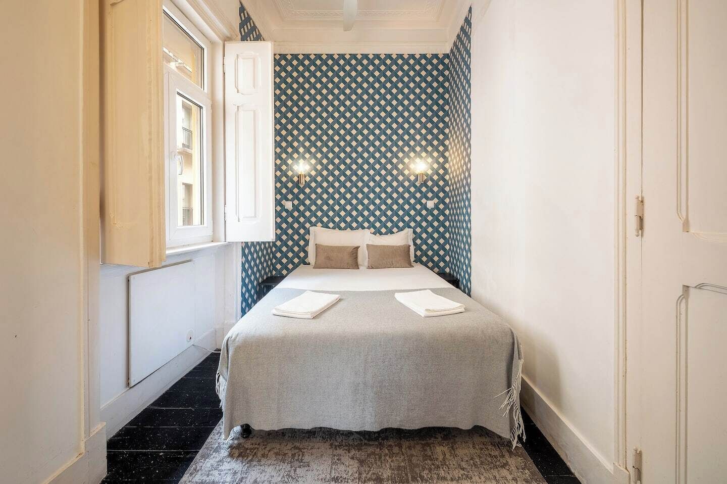 Kapea hotellihuone, jossa on sänky, kuvioitu seinä ja pieni ikkuna.