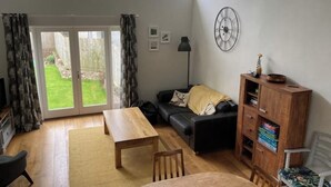 Een woonkamer met een houten tafel, een zwarte bank, een boekenkast en een groot raam met gordijnen.