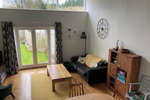 Ein Wohnzimmer mit Holztisch, schwarzem Sofa, Bücherregal und einem großen Fenster mit Vorhängen.