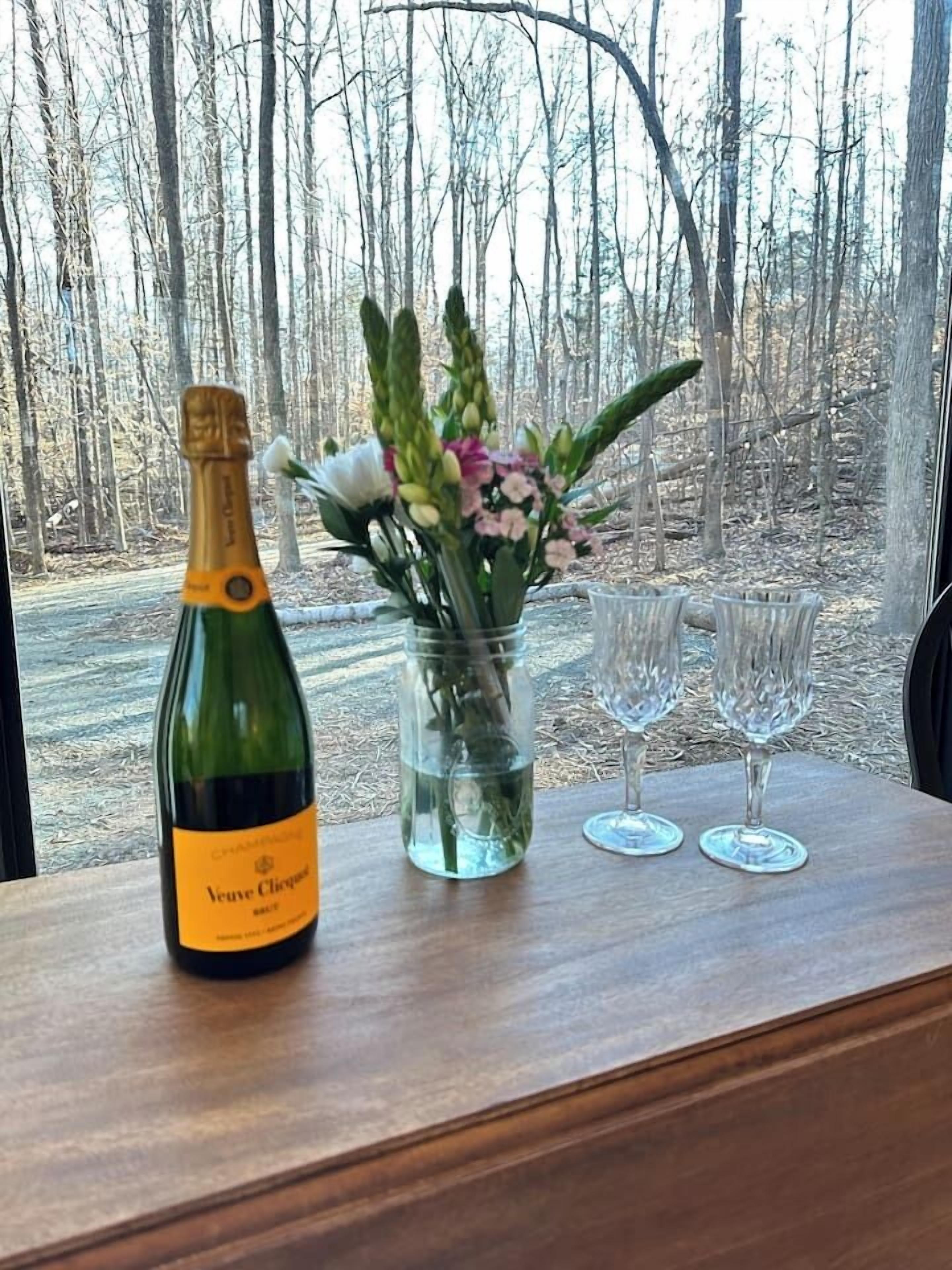 Una botella de Veuve Clicquot champagne, un florero con flores y dos copas sobre una mesa de madera con vista a los árboles.