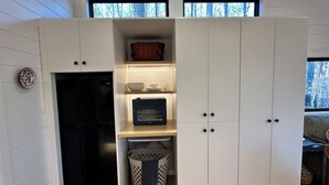 A compact kitchen with a built-in refrigerator, a microwave, and a small shelf with dishes. - Riverside Studio | Trails, Views, Firepit, Hammock (Elon)