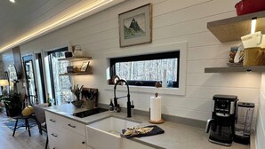 A modern kitchen with a large window, white cabinetry, and a coffee maker. - Riverside Studio | Trails, Views, Firepit, Hammock (Elon)