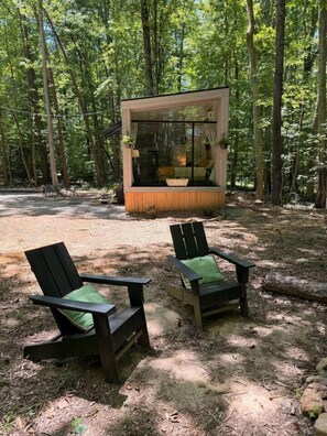 A cabin with a glass front, surrounded by a forest. There are two Adirondack chairs with green cushions. - Riverside Studio | Trails, Views, Firepit, Hammock (Elon)