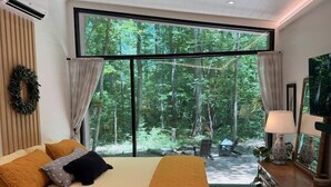 A bedroom with a bed, a desk, a chair, a TV, and a large window with a view of trees. - Riverside Studio | Trails, Views, Firepit, Hammock (Elon)