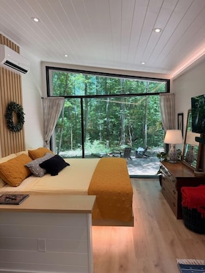A bedroom with a bed, a desk, a chair, a TV, and a large window with a view of trees.