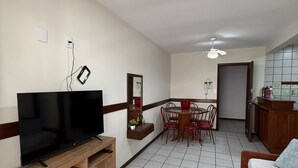 A modern living room with a flat-screen TV on a wooden stand, a dining area with a table and chairs, and a kitchenette with cabinets and a countertop. - Viva Canasvieiras Apartment 50m from the beach (Florianópolis)