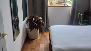 Ein Schlafzimmer mit einem Bett, einer Zimmerpflanze, einem Spiegel und einem Fenster mit Vorhängen.
