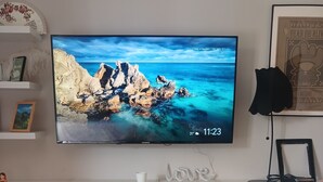 Ein Fernsehgerät zeigt eine malerische Küstenansicht, an der Wand montiert, mit einem weißen Regal darunter, das verschiedene Gegenstände beherbergt.