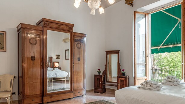 A bedroom with a bed, a wooden wardrobe, a mirror, a window with a green curtain, and a chair. - Beautiful and spacious modern apartment in Florence, located in the quiet and well-connected Campo di Marte area. Situated on the first floor, the apartment can accommodate up to 7 guests with 2 double beds and 3 single bed, arranged across 4 bedrooms. I (Firenze)