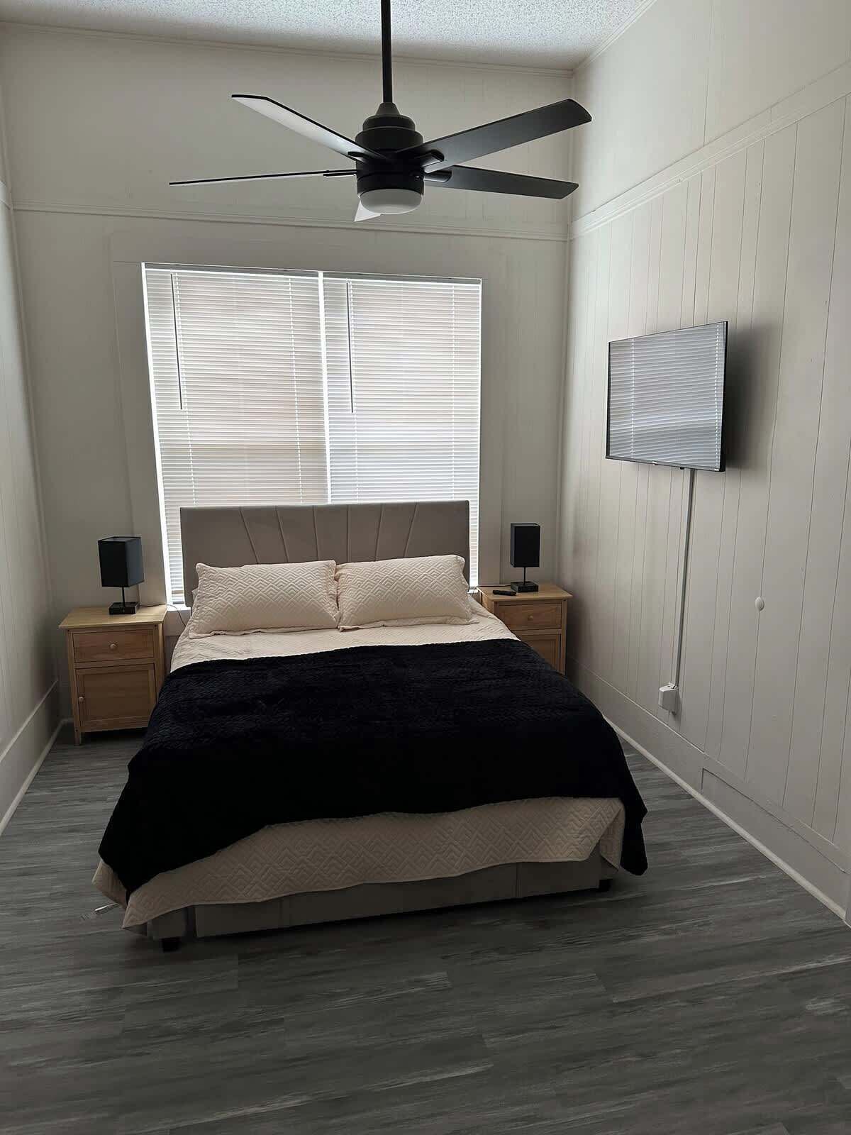 Un dormitorio con cama, ventilador de techo, televisor montado en la pared y ventana con persianas.
