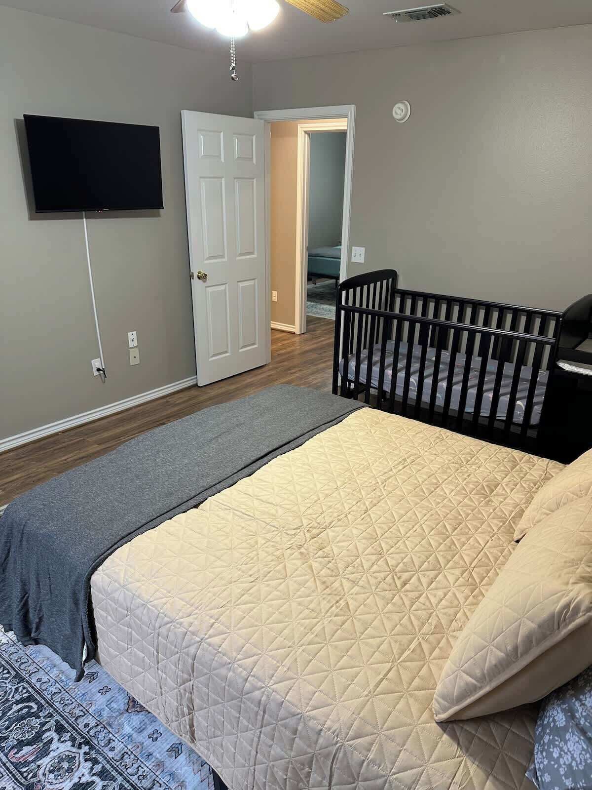 A bedroom with a bed, a crib, a television mounted on the wall, and a door leading to another room.