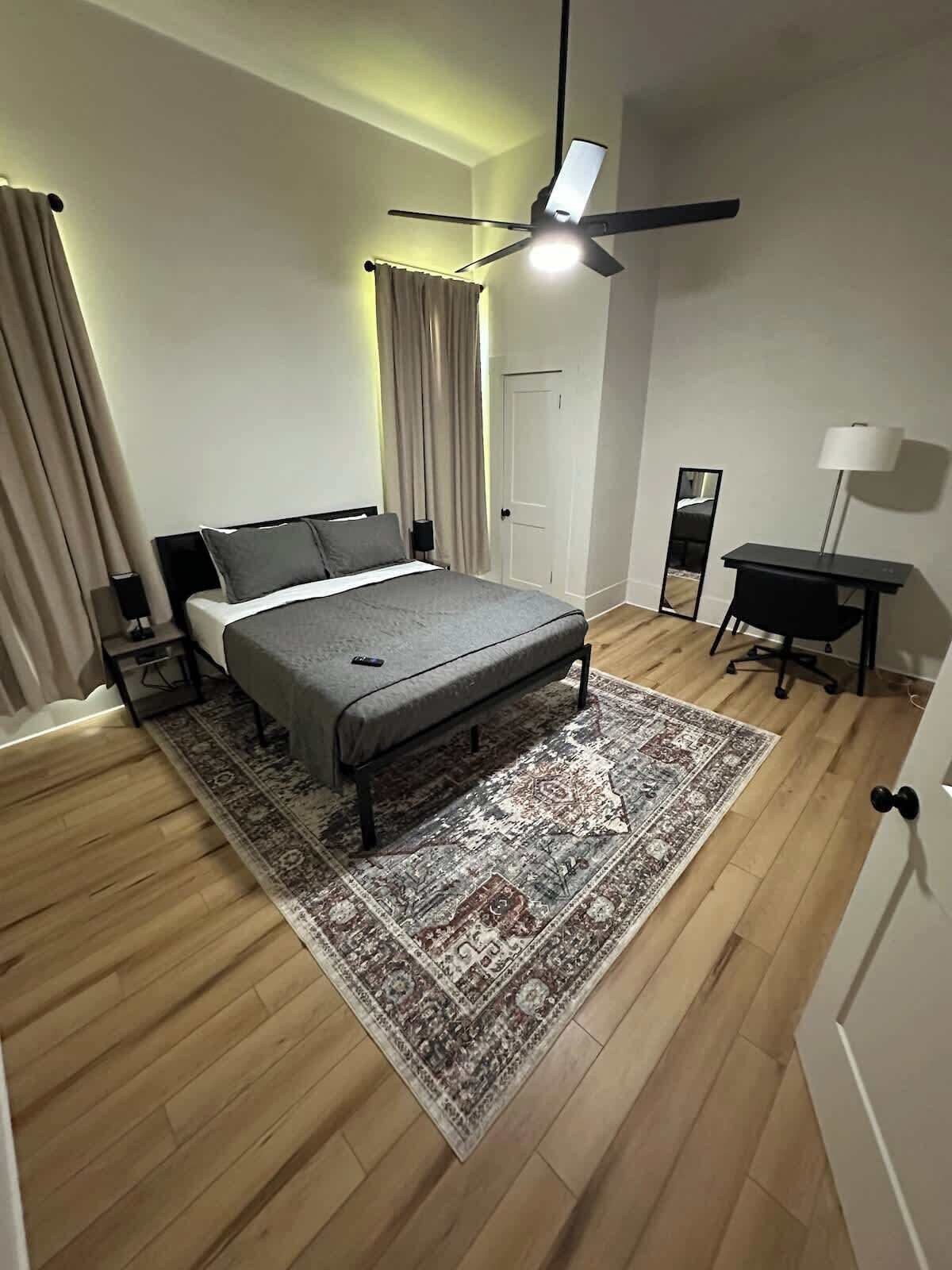 A modern bedroom with a bed, a desk, a chair, a standing lamp, a ceiling fan, and a large rug.