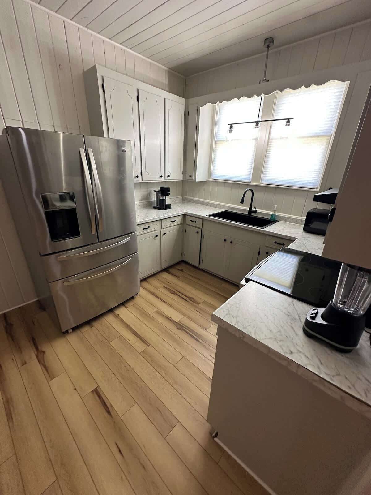 A modern kitchen with white cabinets, a stainless steel refrigerator, a built-in oven, a sink, and a window with blinds.