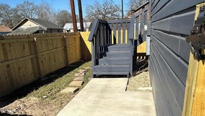 Un camino de concreto que lleva a una escalera de madera con barandilla, rodeado por una valla de madera y un edificio con fachada oscura.