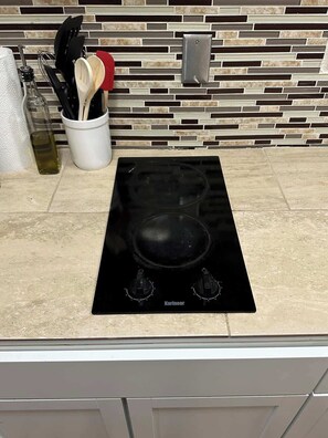 A kitchen with a black induction cooktop, a tiled backsplash, and a countertop with various utensils and a bottle of olive oil.