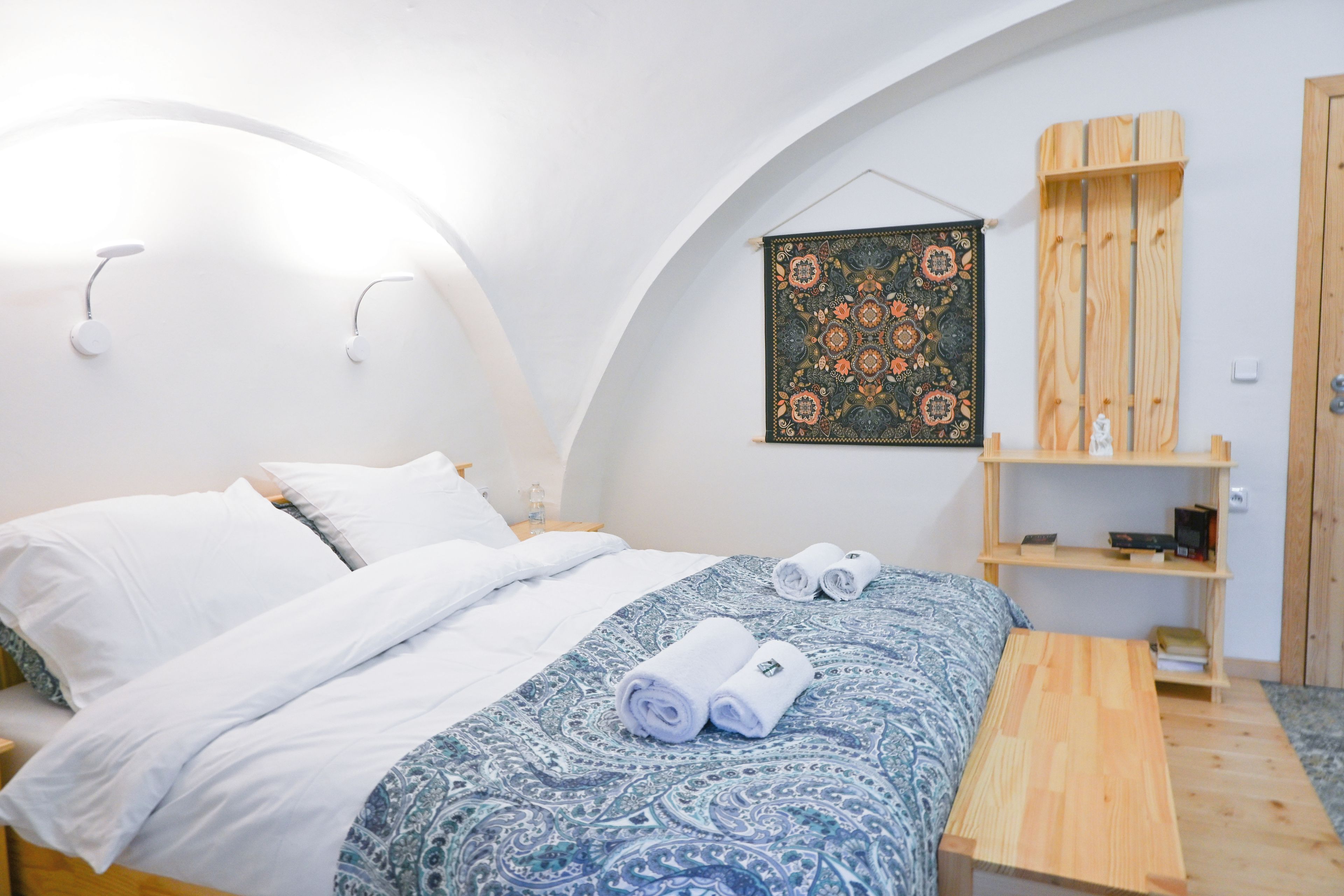 A bedroom with a bed, pillows, a bedside table, a wall-mounted artwork, and a wooden shelf.