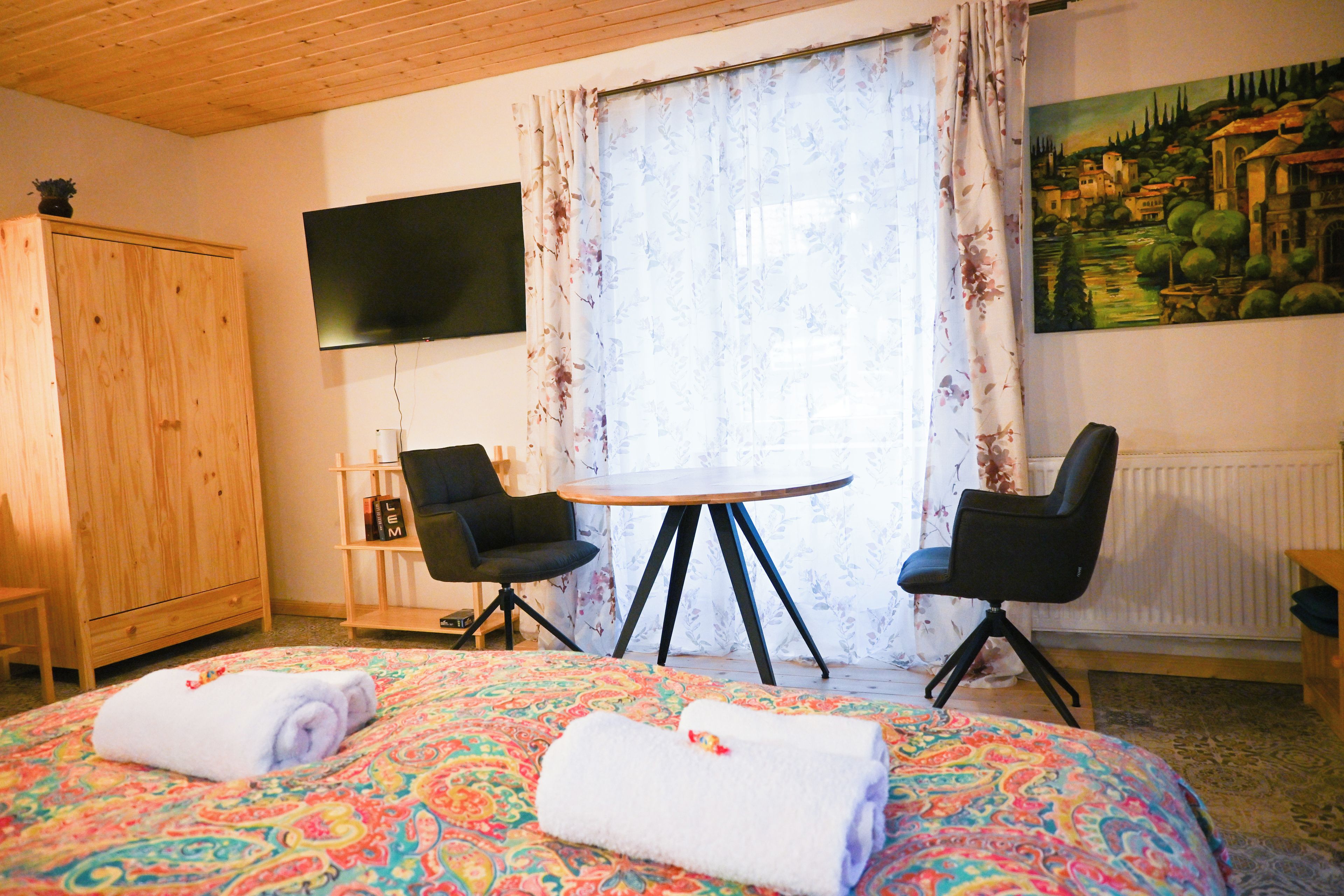 A hotel room with a bed, two chairs, a small table, a TV, and a painting on the wall.