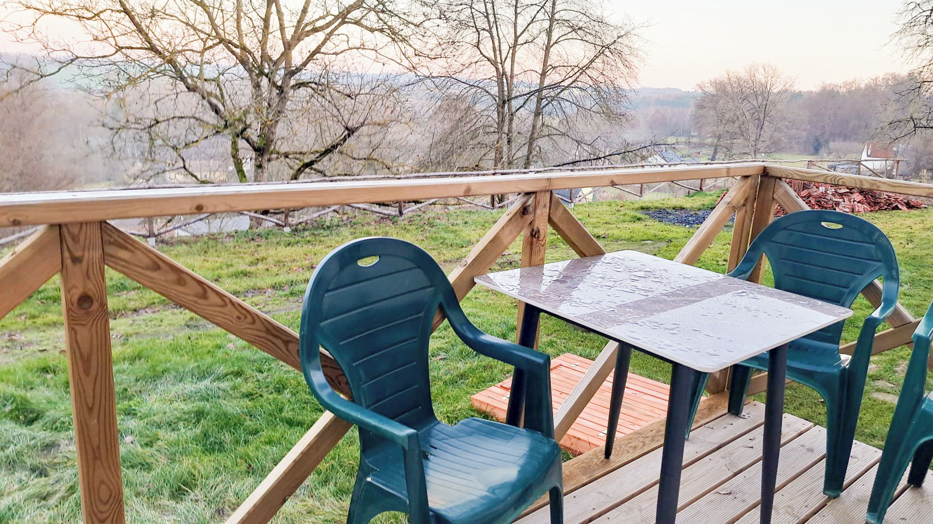 Una terraza al aire libre con una mesa y sillas, rodeada por barandilla de madera, con vista a un área verde con árboles.