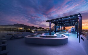 Ett poolområde på taket med en överbyggd sittplats, en pool med en unik design och utsikt över stadsbilden i skymningen.