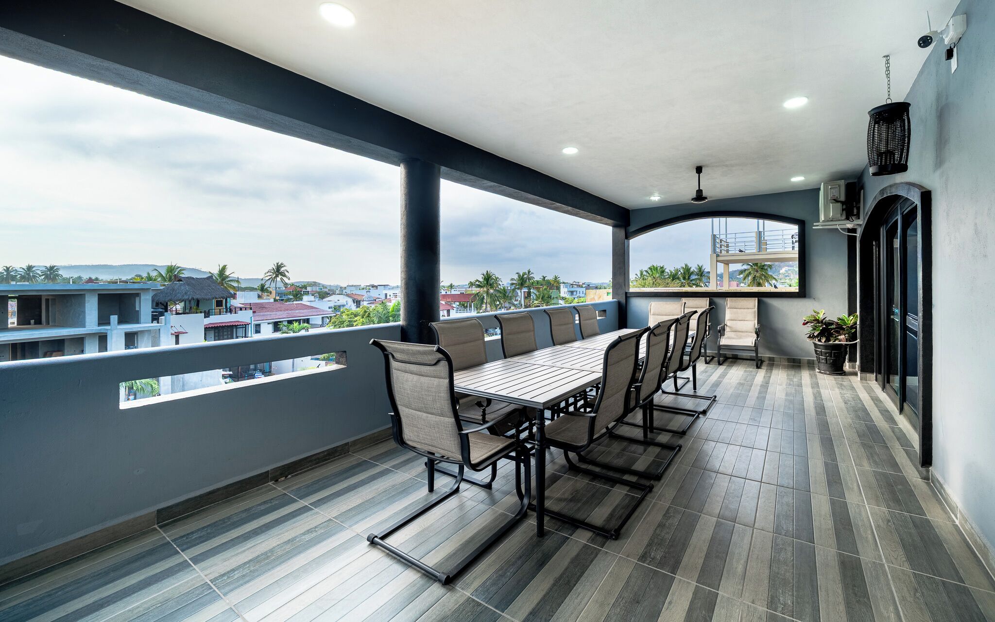 Ein Balkon mit Esstisch und Stühlen, einer hängenden Leuchte und Blick auf Gebäude und Palmen.