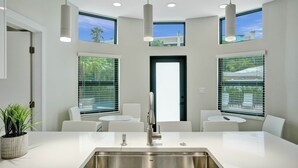 A modern kitchen with a stainless steel sink, white countertops, and pendant lighting. There are two white chairs and a small table. - Deerfield Luxury Stay: Heated Pool, Hot Tub & Golf (Deerfield Beach)