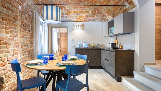 A dining area with a brick wall, a wooden table, blue chairs, and a hanging light fixture. - Hostdomus - The Court (Alba)