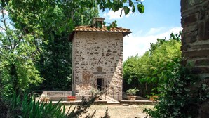 Um edifício de pedra com telhado em tijolos, cercado por vegetação e um espaço pavimentado.
