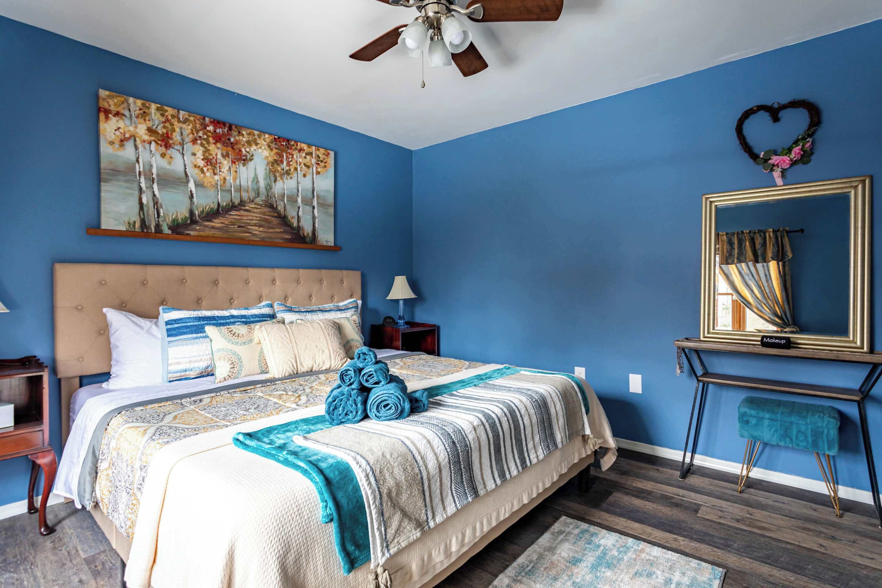 Een slaapkamer met een groot bed, een plafondventilator, een schilderij van bomen, een spiegel en een decoratie in de vorm van een hart aan de muur.