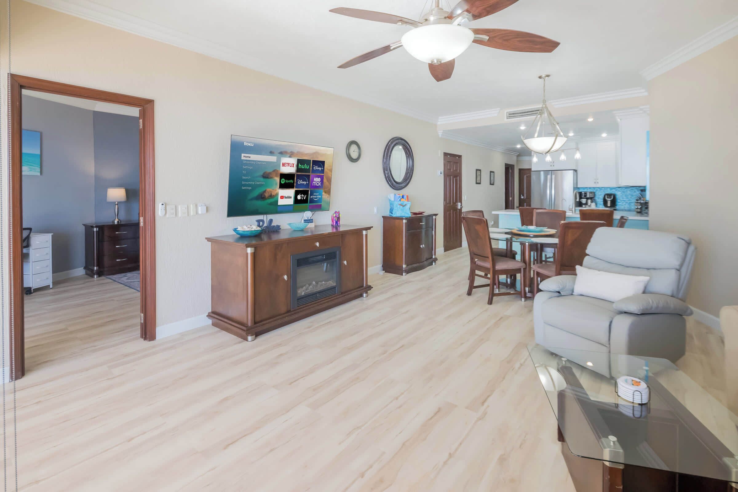 A modern living room with a fireplace, a television displaying streaming services, a dining area with a table and chairs, and a kitchen in the background.