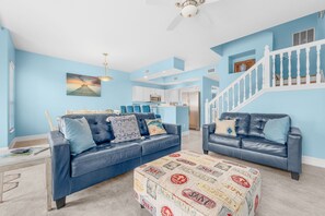 A living room with two blue leather sofas, a coffee table with surf-themed designs, a ceiling fan, and a staircase with white railings. - Pool, Steps to European Village & Trails (Palm Coast)