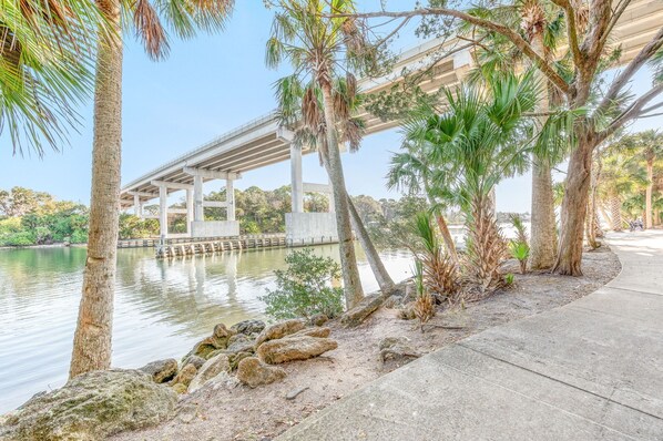 A concrete walkway beside a calm body of water, with palm trees and a bridge in the background.