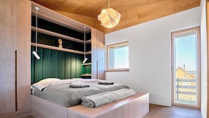 Ein modernes Schlafzimmer mit Bett, eingebauten Stauraumlösungen, einem großen Fenster und Blick auf ein Haus.
